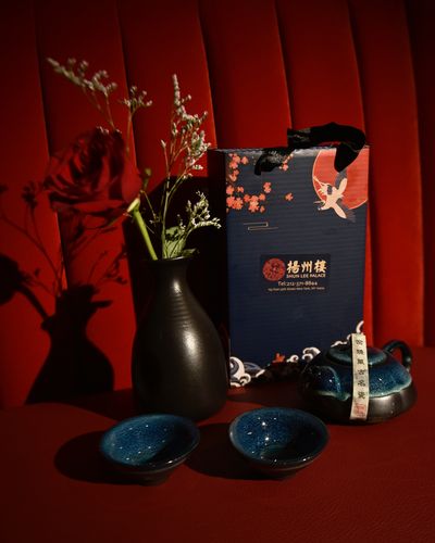 A navy Shun Lee Palace 扬州楼 gift bag with a teapot, teacups and white flowers on a red velvet banquette