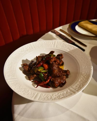 Orange beef with dried red chilies and green peppers in an embossed white bowl at the edge of a set table, with a red velvet banquette behind