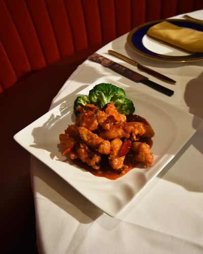 General Tso's chicken with dried chilies and broccoli on a square white plate at the edge of a set table, with a red velvet banquette behind