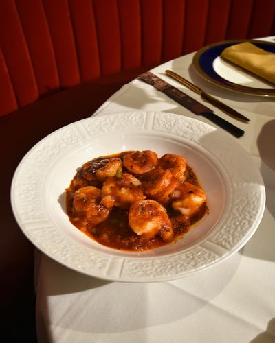 Szechuan prawns in a glossy red chili sauce in an embossed white bowl at the edge of a set table, with a red velvet banquette behind