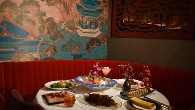A round table set with a spread of dishes (a whole crispy fish, a blue-rimmed sashimi bowl with orchids, prawns with broccoli, tuna on a pink salt block, a sushi roll and cocktails) in front of a red velvet banquette, a painted palace mural with a pagoda and a carved wooden screen