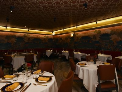 The main dining room: round tables with white cloths, navy-and-gold chargers and gold napkins, under a painted palace mural that runs around the room below a gold-lit ceiling cove and patterned ceiling