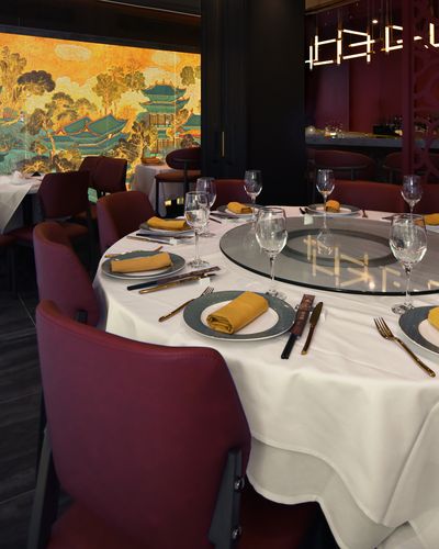 A large round banquet table set for a party with a glass lazy susan, wine glasses, gold napkins and navy-rimmed plates, in front of a gold-and-teal painted palace mural and a red carved screen