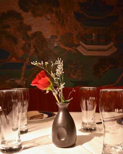 A red rose and dried flowers in a matte black vase on a white tablecloth, between water glasses etched with the 扬州楼 Shun Lee Palace name, with the painted palace mural softly lit behind