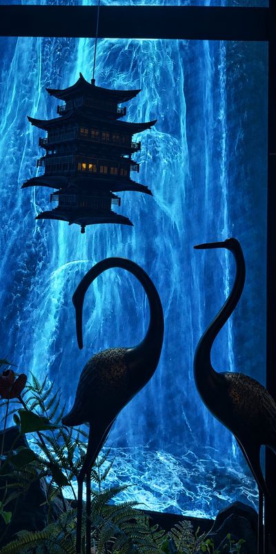 The entrance water wall: a blue-lit cascade behind a bronze pagoda and silhouetted cranes among ferns.
