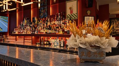 The bar: a marble counter with a golden wheat arrangement in a “生意兴隆” planter, before a red wall lined with spirits under white linear lights.