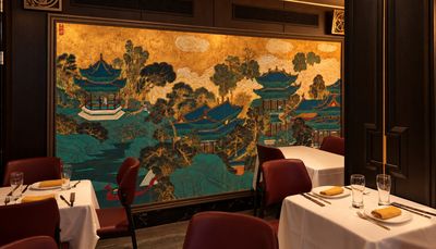 A small dining room beside a large gold-and-teal painted mural of palaces among pine trees, tables set in white linen.