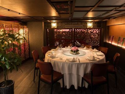 A private dining room: a round table for ten in white linen with a glass lazy susan and pink flowers, before a red cloud-pattern carved screen, with a glowing lattice screen and a money tree to the left.